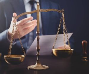 Une personne en costume tient un document , avec une balance de justice en laiton au premier plan sur une surface sombre.